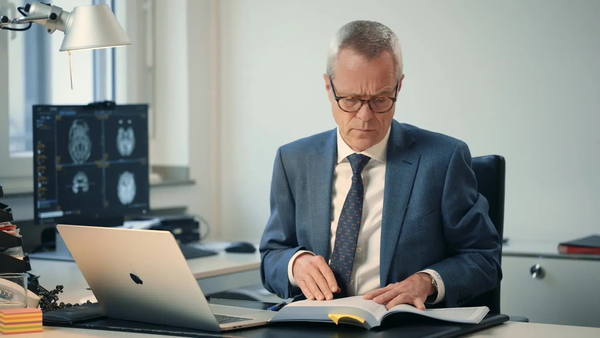 Neurowissenschaftler am Schreibtisch mit Laptop und Fachbuch, im Hintergrund MRT-Aufnahmen — Szene aus dem DFG-Implicit-Bias-Film